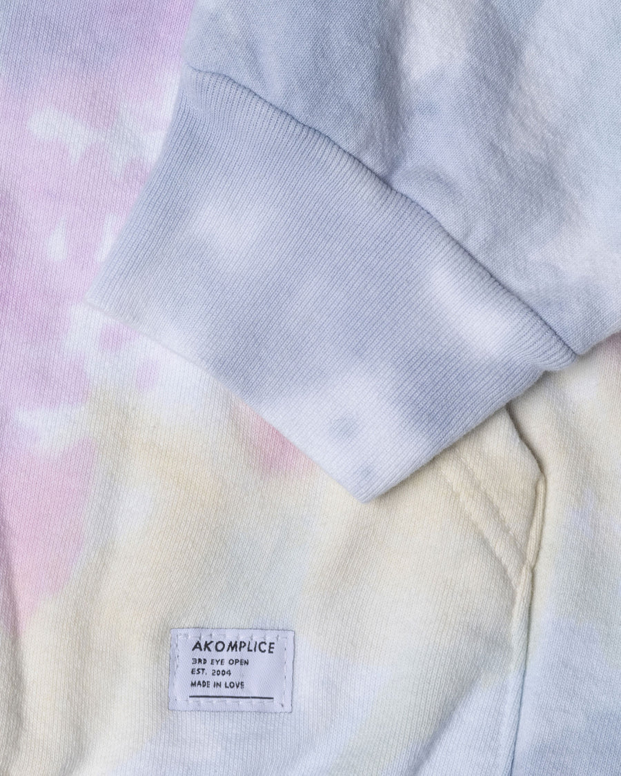 Close-up of a hoodie with a tie-dye pattern and Akomplice label on a pink and yellow gradient background.