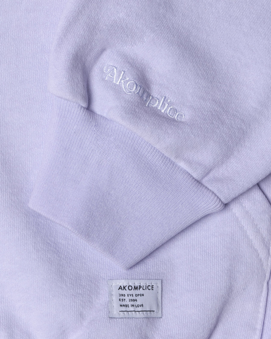 Close-up of a lavender hoodie with 'AKOMPLICE' branding on the chest pocket and label.