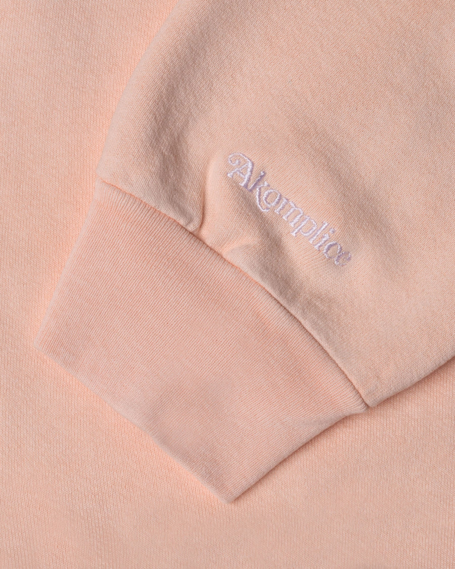 Close-up of a pink sweatshirt with a brand logo on the sleeve against a matching background.