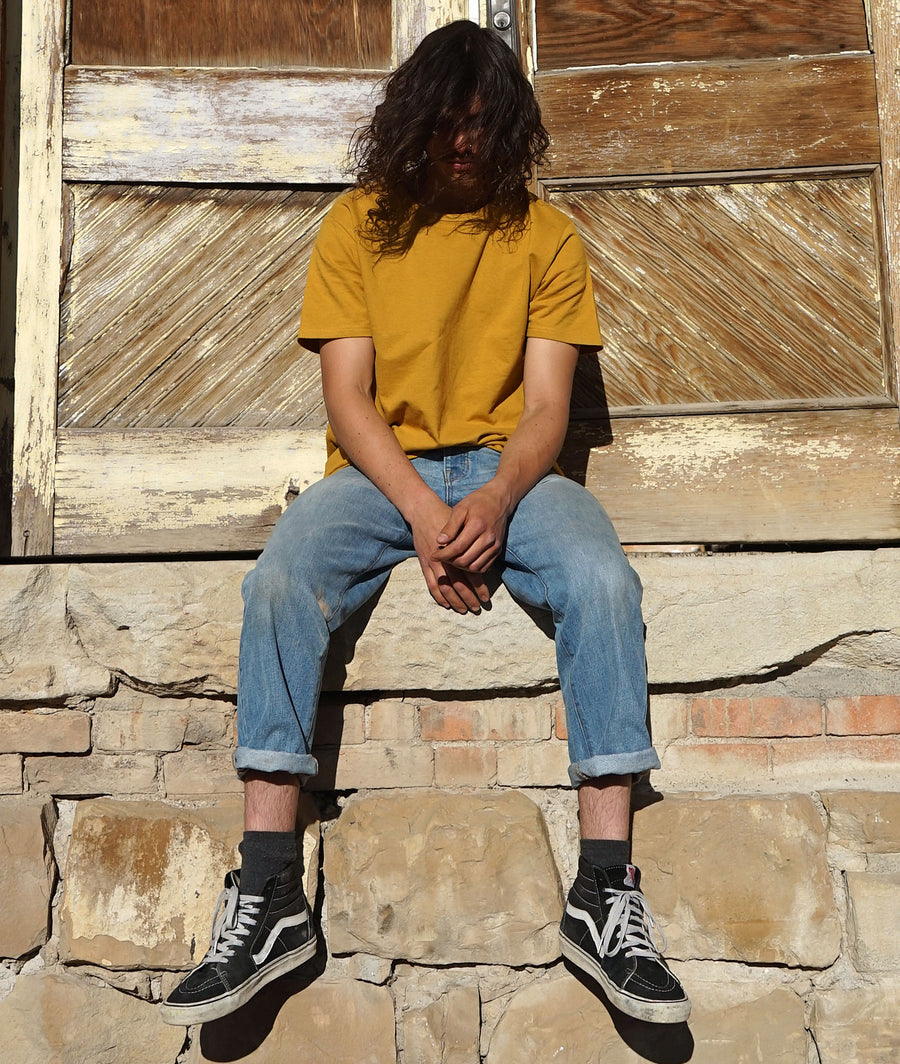 Person wearing a mustard yellow shirt, blue jeans, and black sneakers sitting on stone steps with a wooden door in the background.