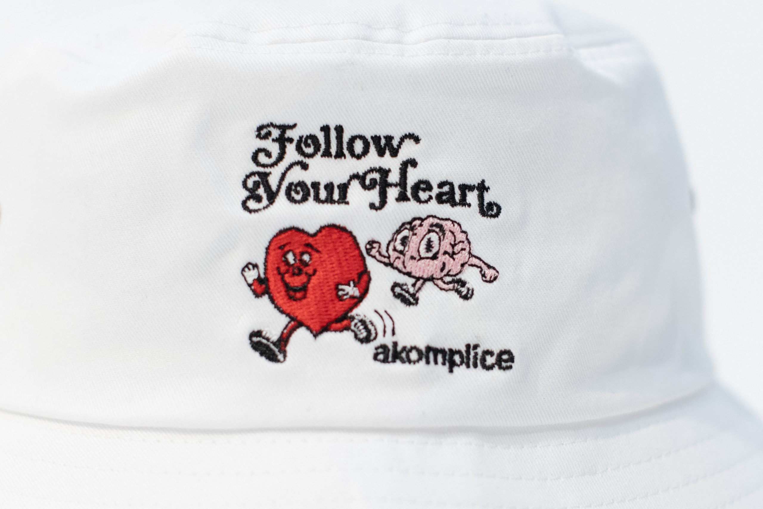 White cap with embroidered design of a heart and brain running together, text 'Follow Your Heart', and brand 'akomplice'.