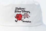 White cap with embroidered design of a heart and brain running together, text 'Follow Your Heart', and brand 'akomplice'.